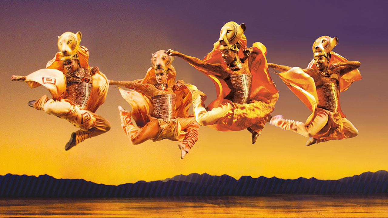 Sehen Sie spektakuläre Bühnenbilder und Kostüme. Photo: Johan Persson. Bild mit Genehmigung von Disney The Lion King / Urheberrecht Johan Persson. Vier Tänzerinnen und Tänzer tanzen in Löwenkostümen in Der König der Löwen über die Bühne.