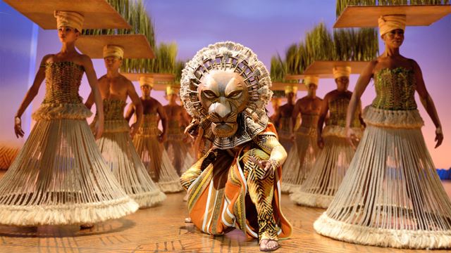 Le personnage de Mufasa se tient au centre de la scène et est antourée d'un clan de lionnes.