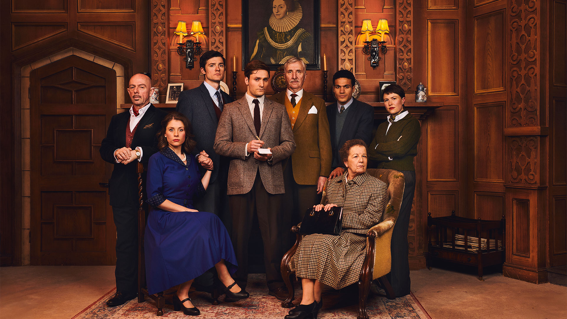 The cast of The Mousetrap sit and stand on stage, looking mysteriously at the camera. Two women sit down, and the men stand behind them.