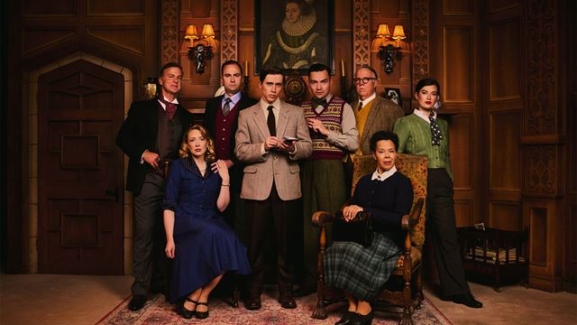 The cast of The Mousetrap sit and stand on stage, looking mysteriously at the camera. Two women sit down, and the men stand behind them.