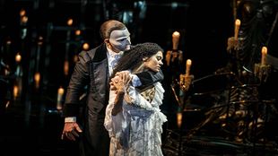 A man wearing a white mask covering half its face is holding a woman on the stage of Phantom of the Opera with many candles illuminating the stage.