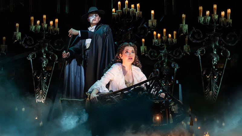 The Phantom of the Opera, wearing a mask hiding half of his face, stands above Christine, a young opera singer.