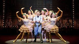 A lively ensemble scene from The Producers musical, with a man in suspenders surrounded by dancers in glittering gold dresses performing under stage lights.