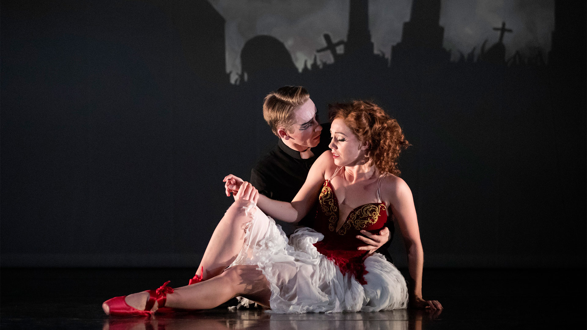 The Olivier Award-winning production of Matthew Bourne's The Red Shoes is returning to London for the festive season. Photo credit: Johan Persson. A man holds a young woman in his arms. She is wearing a bronze corset, a flowy white skirt and red ballet shoes.