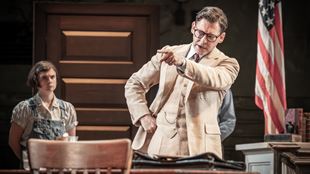 Actor Richard Coyle playing fictional character Atticus Finch on stage, dressed in a beige suit with an American flag in the background.