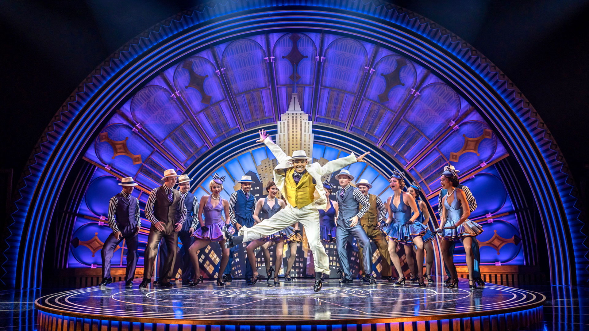 Dressed in a three-piece suit, Phillip Attmore is jumping and dancing on stage with the rest of the Top Hat cast, who are standing at the back.