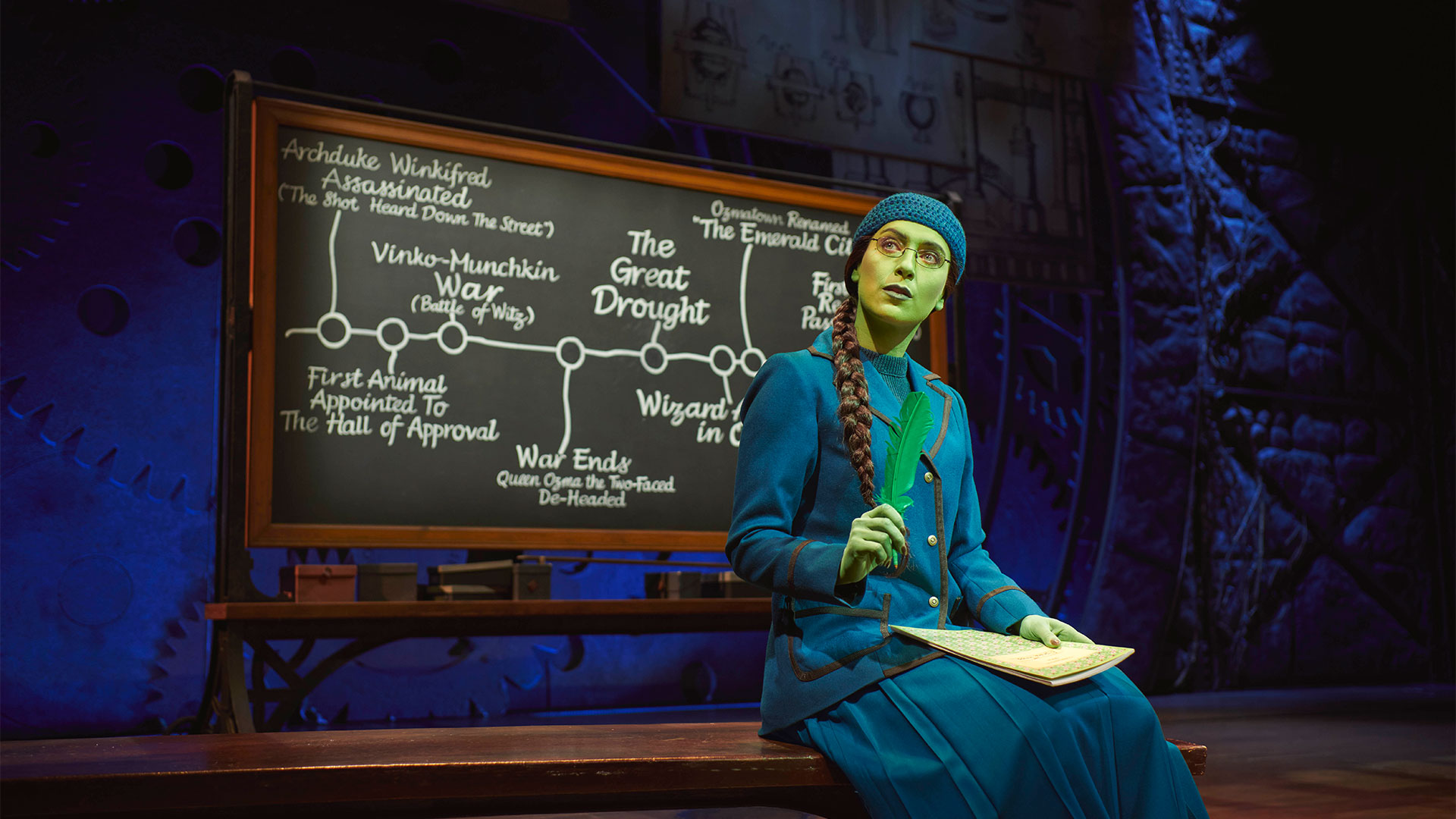 Rediscover the story of the Kingdom of Oz from Elphaba's perspective in Wicked the Musical. Photo credit: Matt Crockett. Elphaba wears the school uniform seating on a bench in front of a black board.