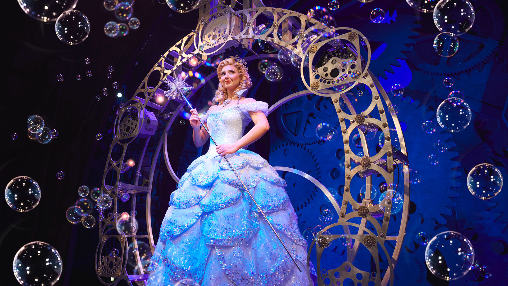 Discover Zizi Strallen playing the role of Glinda the good witch in Wicked the Musical at the Apollo Victoria.Photo credit Matt Crockett. Glinda in Wicked the musical in a blue ball gown in front of a blue and black background with bubbles.