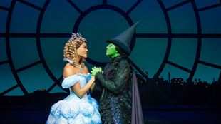 L'actrice qui interprète Glinda dans la comédie musicale Wicked porte une robe de bal bleue et tient la main de l'actrice qui joue le rôle d'Alphaba, célèbre pour sa peau verte, qui porte un chapeau de sorcière et une robe noire.