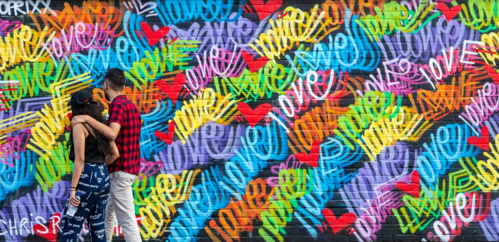 Two people walk arm in arm past a wall of bright rainbow love graffiti.