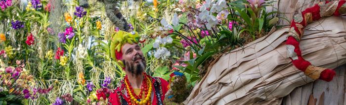A man dressed in colourful clothing and a chameleon on his head tends to a bright orchid display.