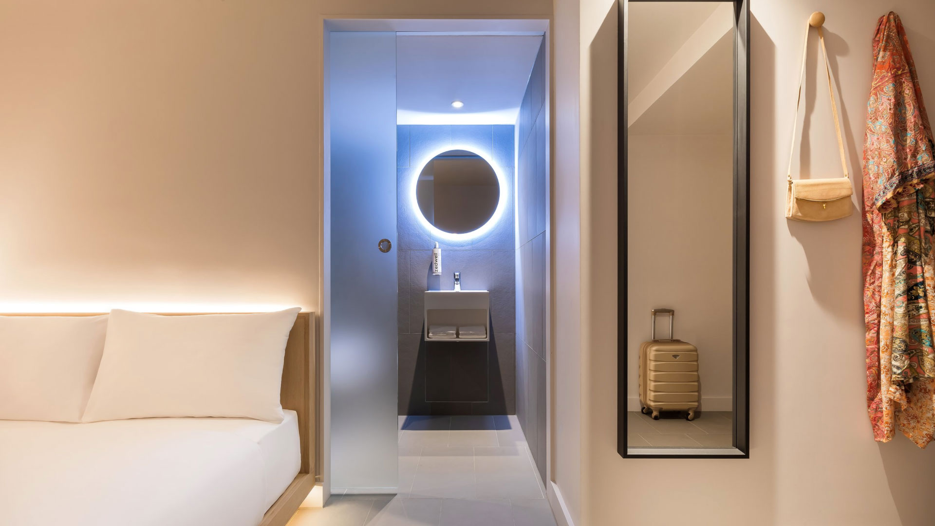 A warm and modern room with wooden-framed double bed, long black mirror and open plan walkway to the bathroom with a light up round mirror