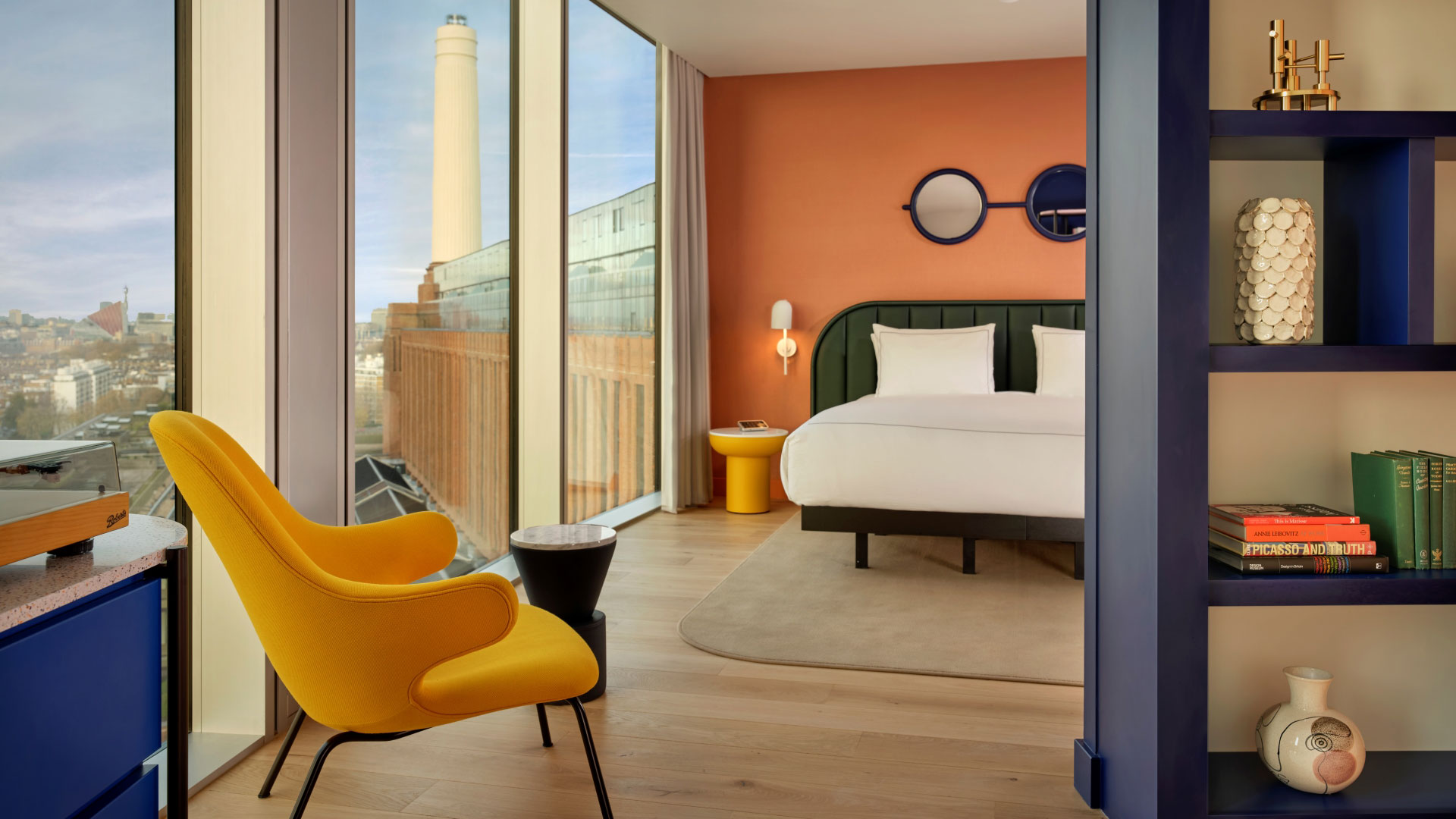 A bedroom filled with colourful furniture and modern artistic touches with floor-to-ceiling windows showing a view of Battersea Power Station in the background