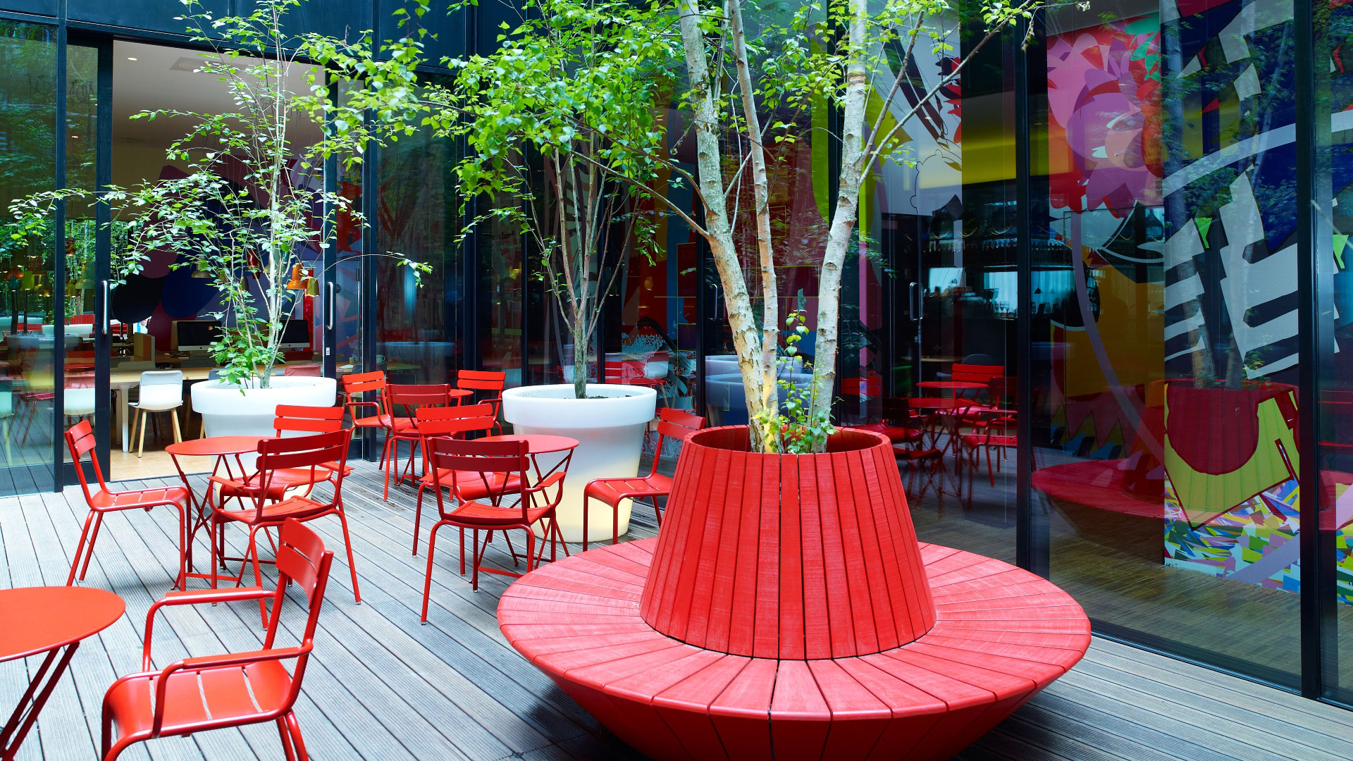 A photo of the outdoor communal space at Citizen M Bankside with red chairs and tables to sit and relax and trees lining the area with floor-to-ceiling windows looking inside the hotel