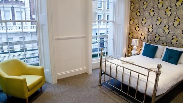 A double bed with white linen and blue cushions, with statement wallpaper behind and a pale green chair in the corner facing the bed.