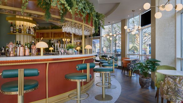 The bar at Doubletree by Hilton London - Chelsea, featuring green leather stools, large windows, foliage and lounge chairs.