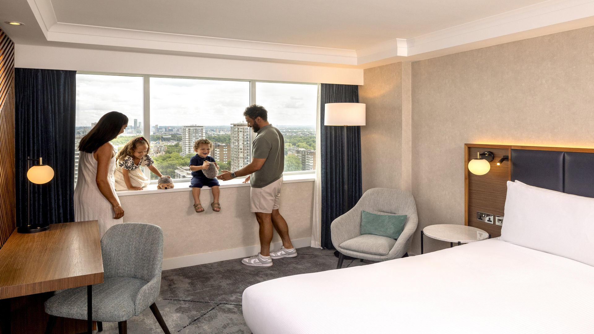 Family of four, two adults and two children sat on the window sill of hotel bedroom, bed and chairs 