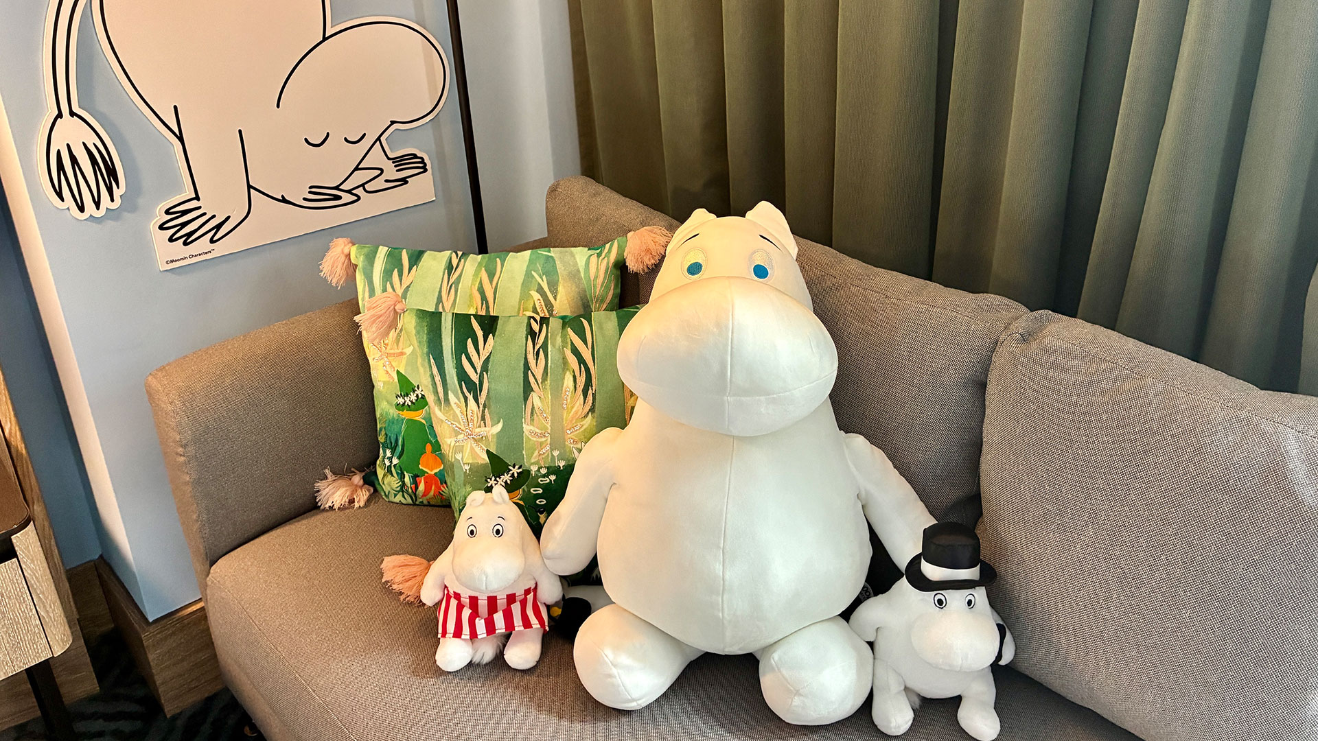 Spend your holidays with the Moomins at Hilton London Bankside. Image courtesy of Chantal Welch. Three Moomin cuddly toys on a grey couch with a Moomin image in the background.