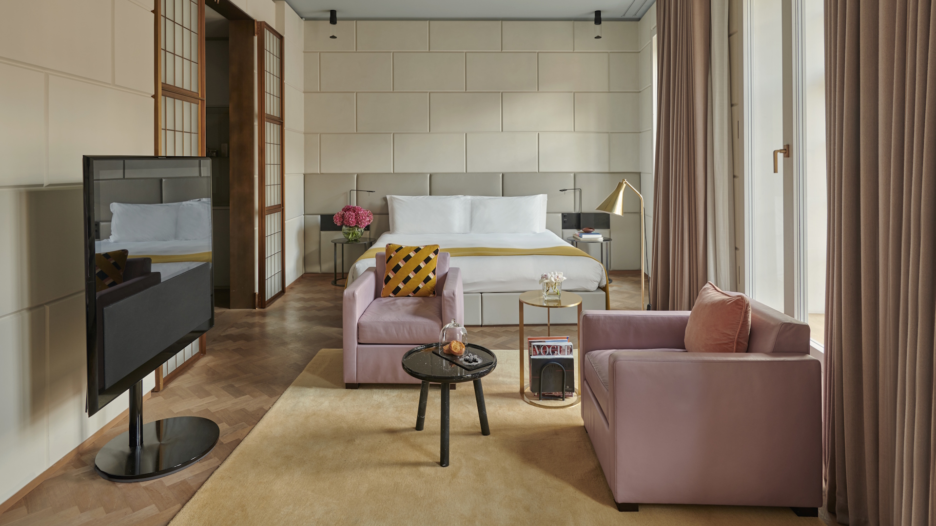 Luxury hotel suite featuring wooden floors, beige walls, white beddings, soft-pink sofa and seats and a huge TV screen.
