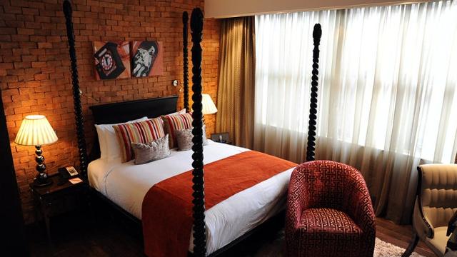 A modern-style four-poster bed up against a bare brick wall with an illuminated lamp on the bedside table.