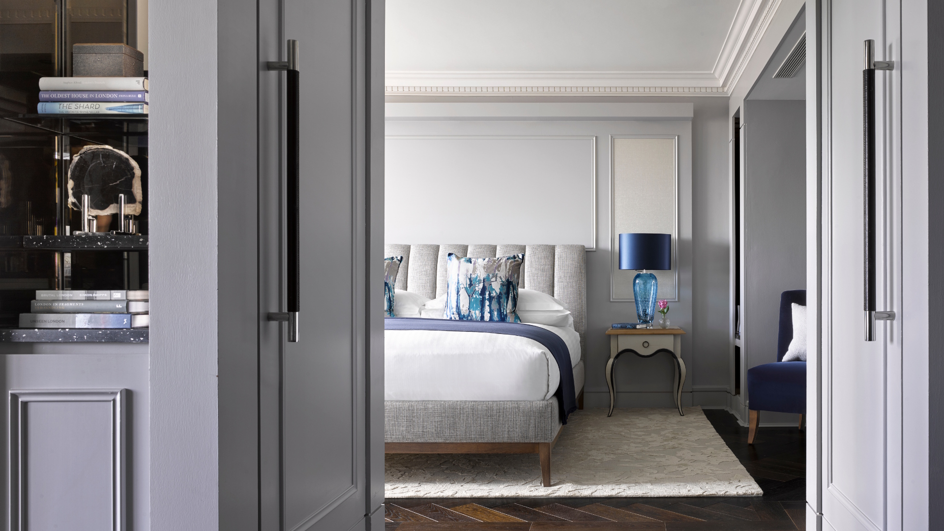 View of a luxury hotel room bed with white linens and blue cushion, with a bedside table and blue bedside lamp.