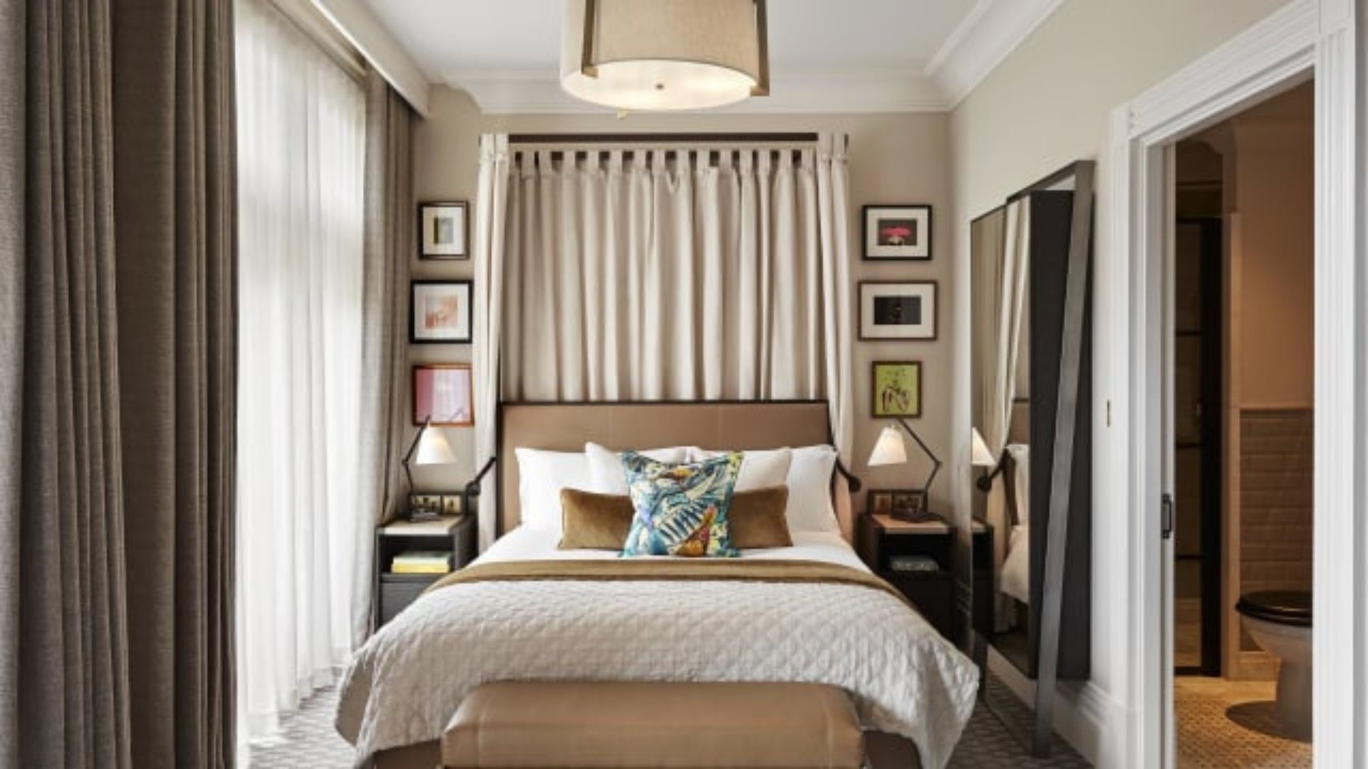 Enjoy a stay at Kimpton Fitzroy London, combining contemporary comfort with grand historic architecture. Image courtesy of Kimpton Fitzroy London. A stylish luxury hotel bedroom at Kimpton Fitzroy London with a king-sized bed, floor-to-ceiling curtains, bedside lamps, framed artwork and an en-suite bathroom entrance.