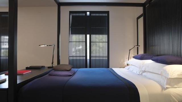 Looking across a double bed towards windows. The bed has a blue duvet and white linen, with a black headboard matching the blinds on the window.