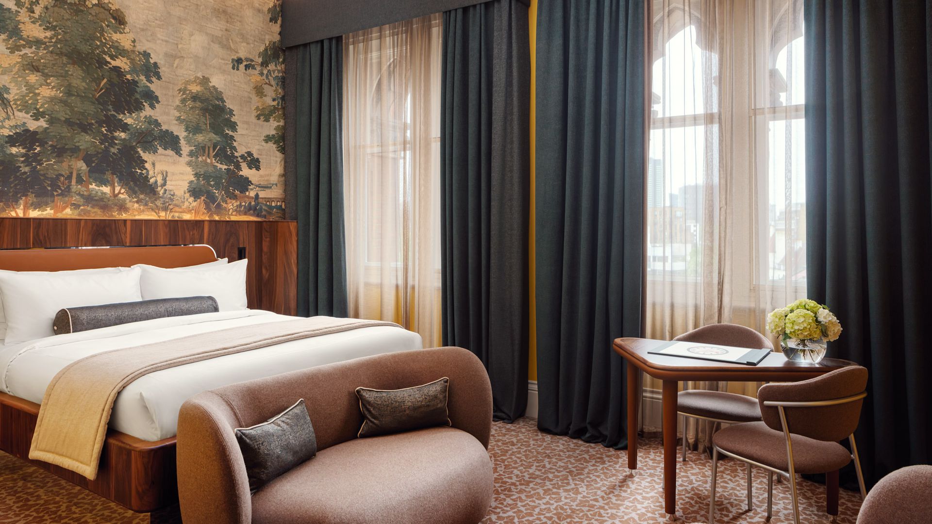 A luxurious guest room at St. Pancras London featuring a king-sized bed, elegant seating area, large arched windows with dark curtains, a mural feature wall and contemporary furnishings.
