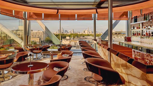 The Emory Rooftop Bar, with stylish modern brown furniture, a long bar and floor-to-ceiling windows in the background showcasing London's skyline.