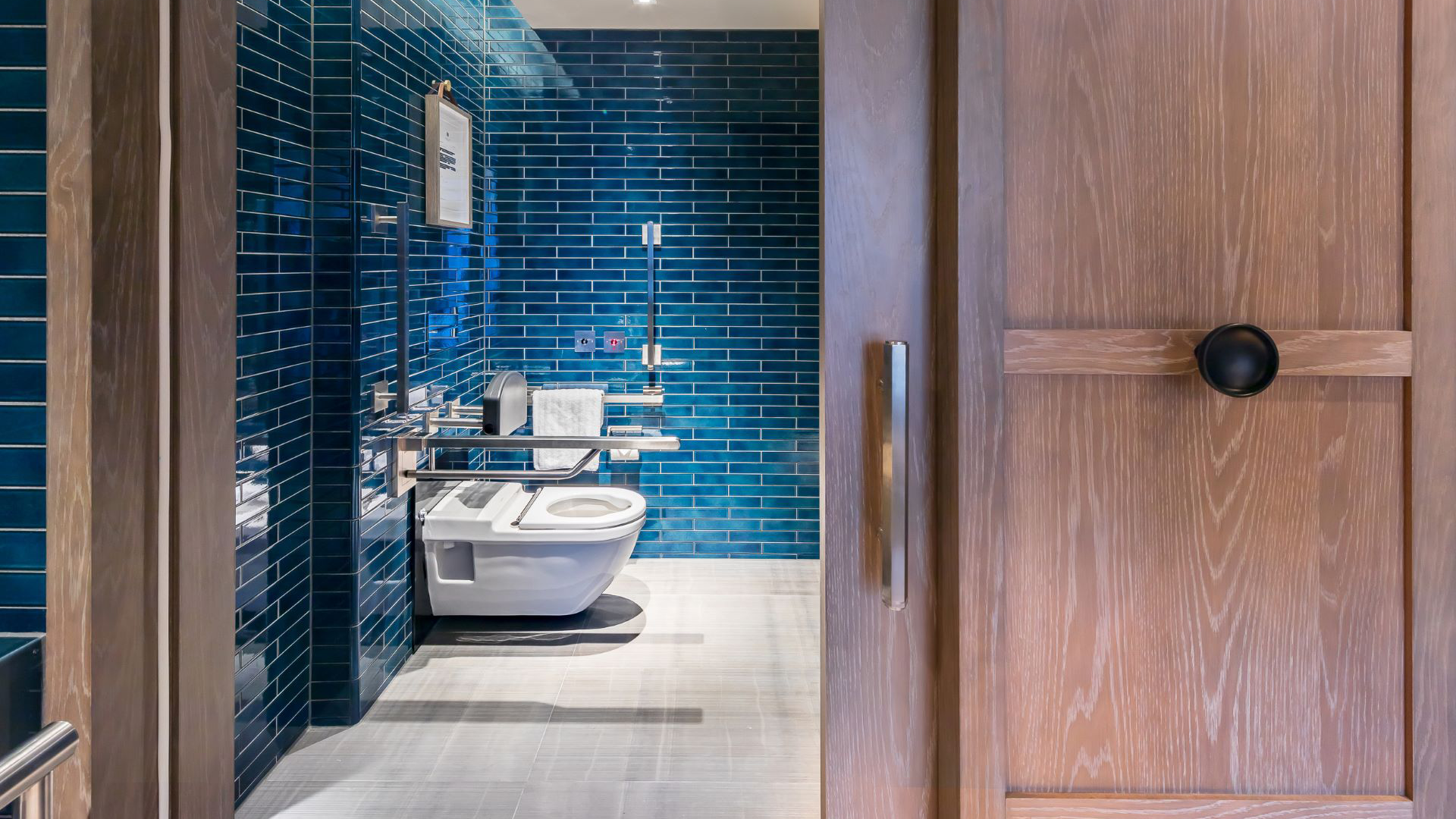 A view of the private bathroom inside an accessible room at The Londoner with modern blue tiles, wooden doors and accessible handrails around the toilet