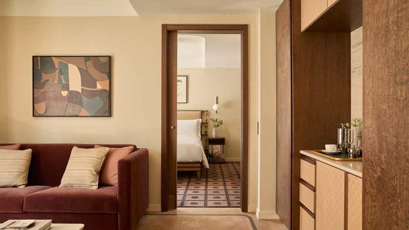 A beautifully designed suite at The Newman Hotel, London, where warm wood tones, soft textures and thoughtful details create a calm, design-led retreat in the heart of the city. Image courtesy of The M Collective. Interior of a stylish hotel suite with a burgundy sofa, wooden kitchenette and an open doorway leading to a serene bedroom, designed in warm neutral and mid-century tones.