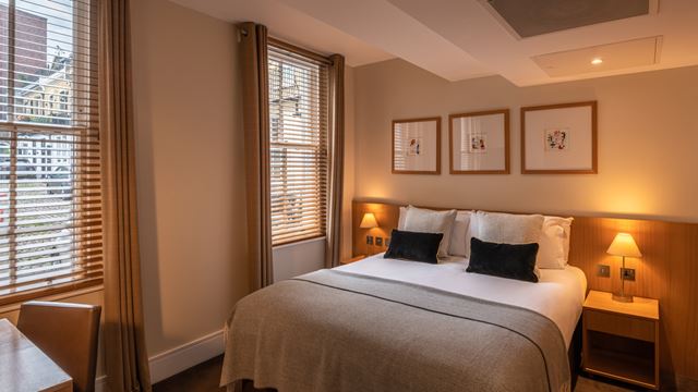 A hotel room decorated in neutral colours with a double bed, side tables either side of the bed and and two large windows next to the bed.