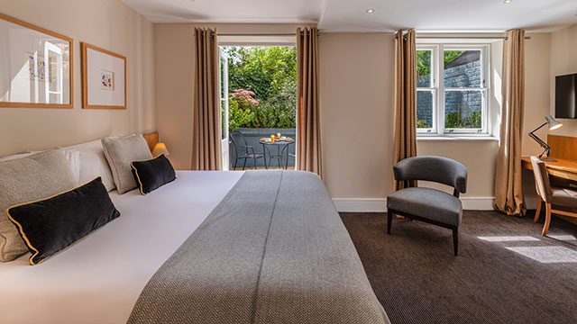 A large bed in the foreground with dark grey cushions and a light grey throw, an armchair and desk on the right and open windows with greenery in the background.