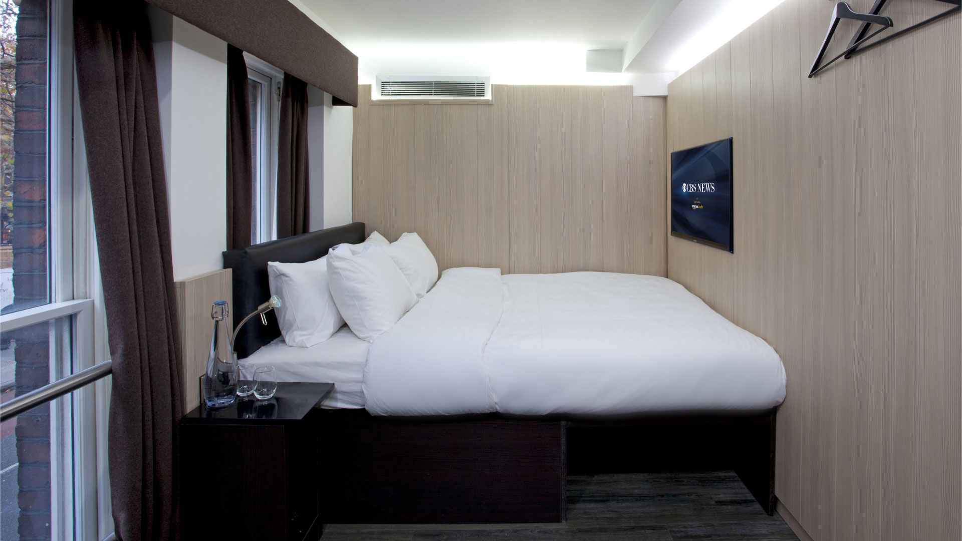 A basic room with a double bed, bedside table, wall-mounted TV and windows