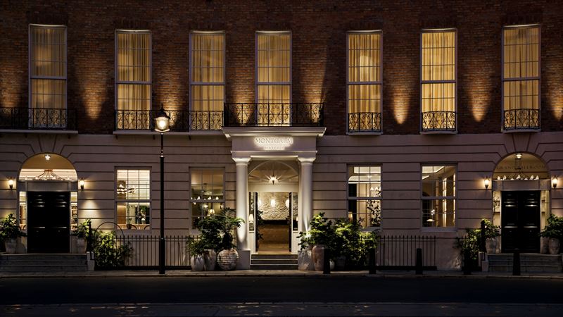 Prepare for a luxurious stay at Montcalm Mayfair situated in one of London's most affluent areas. Credit: Montcalm Mayfair. Image courtesy of Montcalm Mayfair. A photo of the exterior of Montcalm Mayfair hotel at night with warm lighting coming from inside and lighting up the outside of the hotel