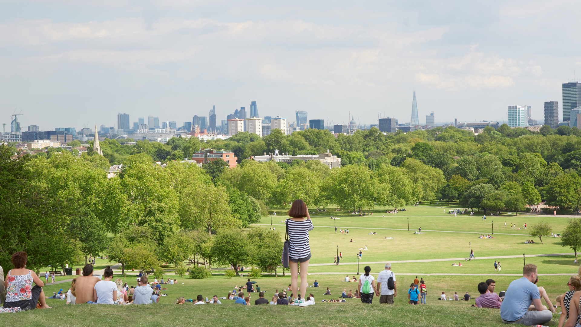A photo of lots of people sat on Primrose Hill facing the London skyline on a sunny summers day with one woman stood up taking a photo